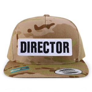 Op de afbeelding: Een camouflage trucker cap met een witte patch waarop in zwarte letters "DIRECTOR" staat. De pet heeft een mesh achterkant en een bruine klep.