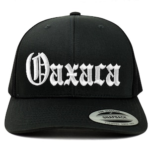 May include: Black trucker hat with a white embroidered "Oaxaca" in a gothic font. The hat has a black mesh back and a black snap closure.