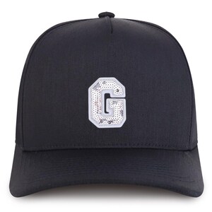 Può includere: Cappellino da baseball nero con una toppa con la lettera "G" bianca e argentata con paillettes sul davanti. Il cappellino ha una visiera curva e un bottone sulla parte superiore. La lettera "G" è bordata di bianco e riempita di paillettes argentate.