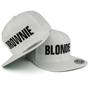 Puede incluir: Dos gorras de béisbol blancas con texto bordado en negro. La gorra de arriba dice "BROWNIE" y la gorra de abajo dice "BLONDIE".