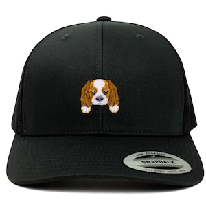 Op de afbeelding: Zwarte truckercap met een geborduurde bruine en witte Cavalier King Charles Spaniel op de voorkant. De pet heeft een zwart mesh achterkant en een zwarte drukknopsluiting. De pet heeft ook een witte sticker op de achterkant met de tekst "The Classics" en "Snapback" met het Yupoong-logo.