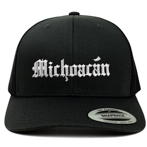 以下が含まれることがあります： 黒いトラッカーハットに、古い英語の文字で「Michoacán」と刺繍された白いデザインが施されています。ハットは黒いメッシュバックと黒いスナップクロージャーが付いています。