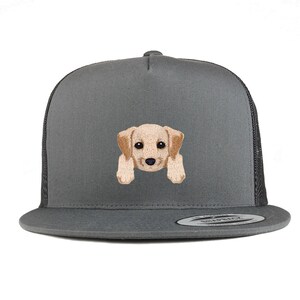 Peut inclure: Un chapeau de camionneur gris et noir avec une image brodée d'un chiot golden retriever. Le chiot regarde la caméra la tête inclinée sur le côté.