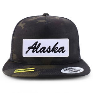 May include: A black and green camouflage trucker hat with a white patch that says "Alaska" in black cursive lettering.