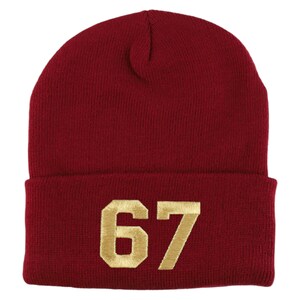 May include: A burgundy knit beanie with a folded brim. The number "67" is embroidered in gold thread on the front. The hat is made of a soft, warm material, suitable for cold weather.