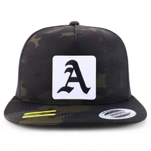 Op de afbeelding: Een zwarte en groene camouflage trucker cap met een witte vierkante patch met een zwarte gestileerde letter 'A'.