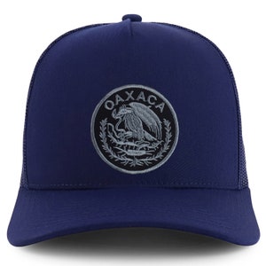 May include: A navy blue trucker hat with a gray embroidered patch featuring the coat of arms of Oaxaca, Mexico. The patch includes the text "OAXACA" around the outer edge.