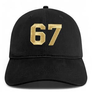 May include: Black baseball cap with the gold number "67" embroidered on the front. The cap has a curved brim and a fabric-covered adjustable strap at the back.