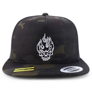 Puede incluir: Gorra snapback negra y con estampado de camuflaje con un diseño de calavera y llamas bordado en blanco. La gorra tiene una visera curva, una parte trasera de malla y un cierre a presión ajustable. Con una etiqueta "MultiCam".