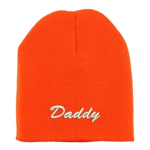 May include: Bright orange beanie hat with the word "Daddy" embroidered in white script. The hat is made of a knitted material and is a solid color. The beanie is a winter accessory.
