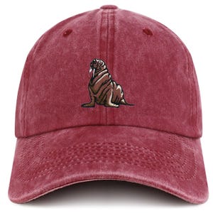 May include: A maroon baseball cap with a distressed finish. The cap features an embroidered walrus design in shades of brown and white. The walrus is sitting upright. The cap has a curved peak and an adjustable strap.