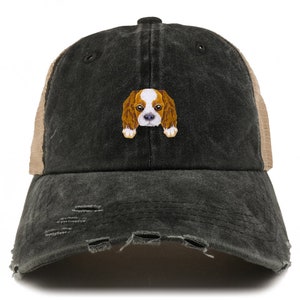 Op de afbeelding: Een zwarte en bruine distressed trucker cap met een klein geborduurd beeld van een Cavalier King Charles Spaniel.