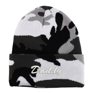 May include: A camouflage-patterned beanie in shades of black, white, and gray. The folded brim features the embroidered word "Daddy" in a cursive font. The hat is made of a soft, knitted material.