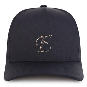 May include: A navy blue baseball cap with a curved brim. The cap features a stylized letter "E" made of small, gold-colored dots on the front. The cap has a button on top.