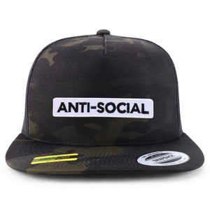 Op de afbeelding: Een zwarte en groene camouflage trucker cap met een witte patch waarop in zwarte letters "ANTI-SOCIAL" staat.