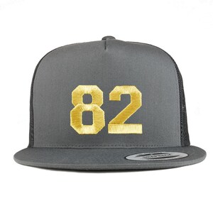 Op de afbeelding: Een grijze truckercap met een zwart mesh achterpaneel. De pet heeft een goudkleurig geborduurd nummer "82" op de voorkant.