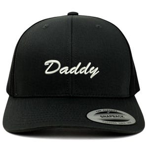 May include: Black baseball cap with the word "Daddy" embroidered in white script across the front. The cap has a mesh back and a snapback closure. The brim is curved, and the cap has a classic, casual style.
