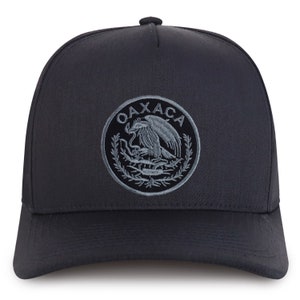 May include: A dark gray baseball cap featuring a circular emblem with the word "OAXACA" and an eagle design. The cap has a structured front and a curved brim, suitable for sun protection.