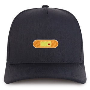 Könnte beinhalten: Eine schwarze Baseballkappe mit einem gelben und orangenen Pflaster mit einem roten Herzen.