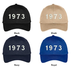May include: Four baseball caps in different colors: black, khaki, navy, and royal blue. Each cap features the year "1973" embroidered in white on the front.