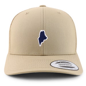 Può includere: Un cappello da camionista beige con un contorno blu e bianco dello stato del Maine sul davanti. Il cappello ha una parte posteriore in rete e una chiusura a scatto. Il cappello ha un'etichetta che dice "The Authentic Yupoong Snapback Yupoong Since 1974".