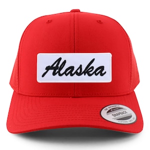 May include: Red mesh trucker hat with a white patch that says "Alaska" in black cursive lettering. The hat has a black "The Authentic Snapback" label on the back.