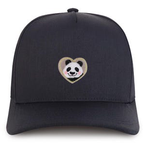 Könnte beinhalten: Marineblaue Baseballkappe mit einem gestickten Panda-Bär-Design in einem herzförmigen Aufnäher. Der Panda hat schwarz-weißes Fell mit einem rosa Rouge. Das Herz ist goldfarben umrandet.