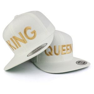 Puede incluir: Dos gorras blancas con texto dorado bordado. Una gorra dice "KING" y la otra "QUEEN". Las gorras tienen una visera plana y una corona estructurada, adecuadas para uso casual.
