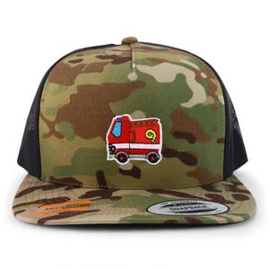 Puede incluir: Gorra snapback con estampado de camuflaje y parte trasera de malla negra. La gorra presenta un parche bordado rojo y amarillo de un camión de bomberos. La visera tiene forma curva y la gorra tiene cierre a presión.