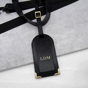 A handmade black personalised leather luggage tag with gold corners on a white and grey stone surface hanging off a black luggage handle