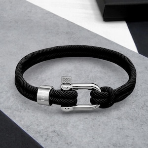 Puede incluir: Pulsera de cuerda negra con cierre plateado. La pulsera presenta un diseño de cuerda gruesa y tejida y un cierre de metal pulido. El cierre tiene un detalle en forma de tornillo y una pieza cilíndrica con las letras "LD" grabadas.