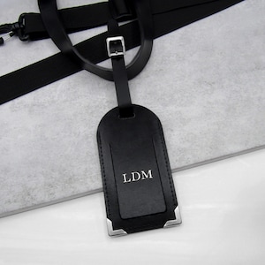 A handmade black personalised leather luggage tag with silver corners on a white and grey stone surface hanging off a black luggage handle