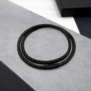 May include: A black braided leather necklace with a magnetic clasp. The necklace is circular and lies on a grey and white surface. The necklace is a jewellery item.