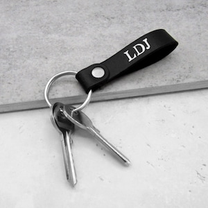 Può includere: Portachiavi in ​​pelle nera con un anello portachiavi argentato e tre chiavi. Il cinturino in pelle ha le iniziali "LDJ" incise in bianco.