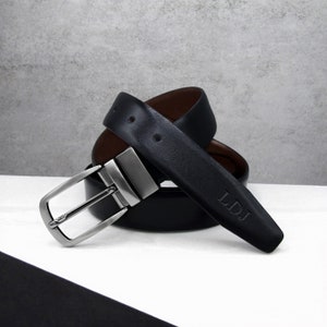 May include: A reversible black leather belt with a silver-tone buckle. The belt has a brown interior and is personalized with the initials "LDJ". The belt is displayed on a white surface.