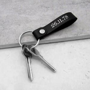 Może przedstawiać: Czarny skórzany brelok do kluczy ze srebrnym kółkiem i trzema kluczami. Pasek skórzany ma wygrawerowaną datę "26.11.78".
