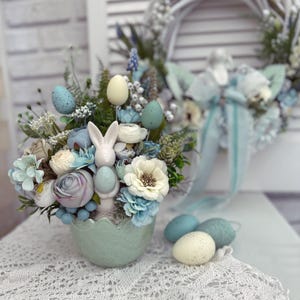 Könnte beinhalten: Ein blauer und weißer Oster-Eiertopf mit einer weißen Hasenfigur im Inneren. Der Topf ist mit blauen und weißen Blumen gefüllt und von blauen und weißen Ostereiern umgeben.