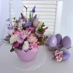 Puede incluir: Arreglo de Pascua en tonos pastel con un jarrón en forma de huevo lila claro lleno de flores artificiales en tonos rosa, morado y blanco. Huevos decorativos y un acento floral completan el tema primaveral.