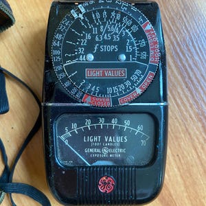 May include: Vintage General Electric exposure meter. The black meter has a dial with f-stops and light values, and a needle gauge. The text "LIGHT VALUES" and "GENERAL ELECTRIC EXPOSURE METER" are visible.