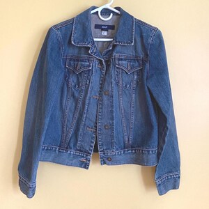 May include: A blue denim jacket with a button-up front, two chest pockets, and a collar. The jacket is hanging on a white hanger. The brand name "GAP" is visible on the label.