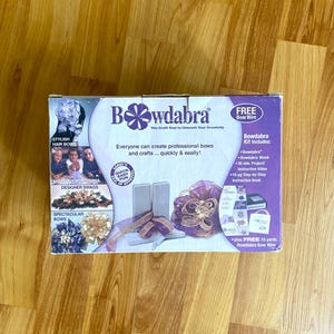 Può includere: Un kit per la creazione di fiocchi Bowdabra su una superficie di legno. La scatola presenta il testo "Bowdabra" e "Tutti possono creare fiocchi e artigianato professionali... rapidamente e facilmente!" Il kit include un Bowdabra, una bacchetta e un libretto di istruzioni.