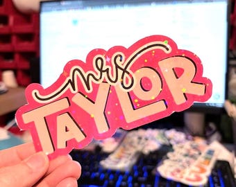 Könnte beinhalten: Ein gestanzter Aufkleber mit dem Text "mrs TAYLOR" in einer fetten, hellen Schriftart mit einer dicken, leuchtend rosa Umrandung. Die Oberfläche des Aufklebers hat ein holografisches Finish mit kleinen, sternförmigen Glitzerpartikeln.