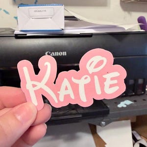 Peut inclure: Un autocollant rose avec le nom "Katie" en lettres blanches. L'autocollant a une bordure blanche et est tenu devant une imprimante noire. D'autres autocollants avec des noms sont visibles.