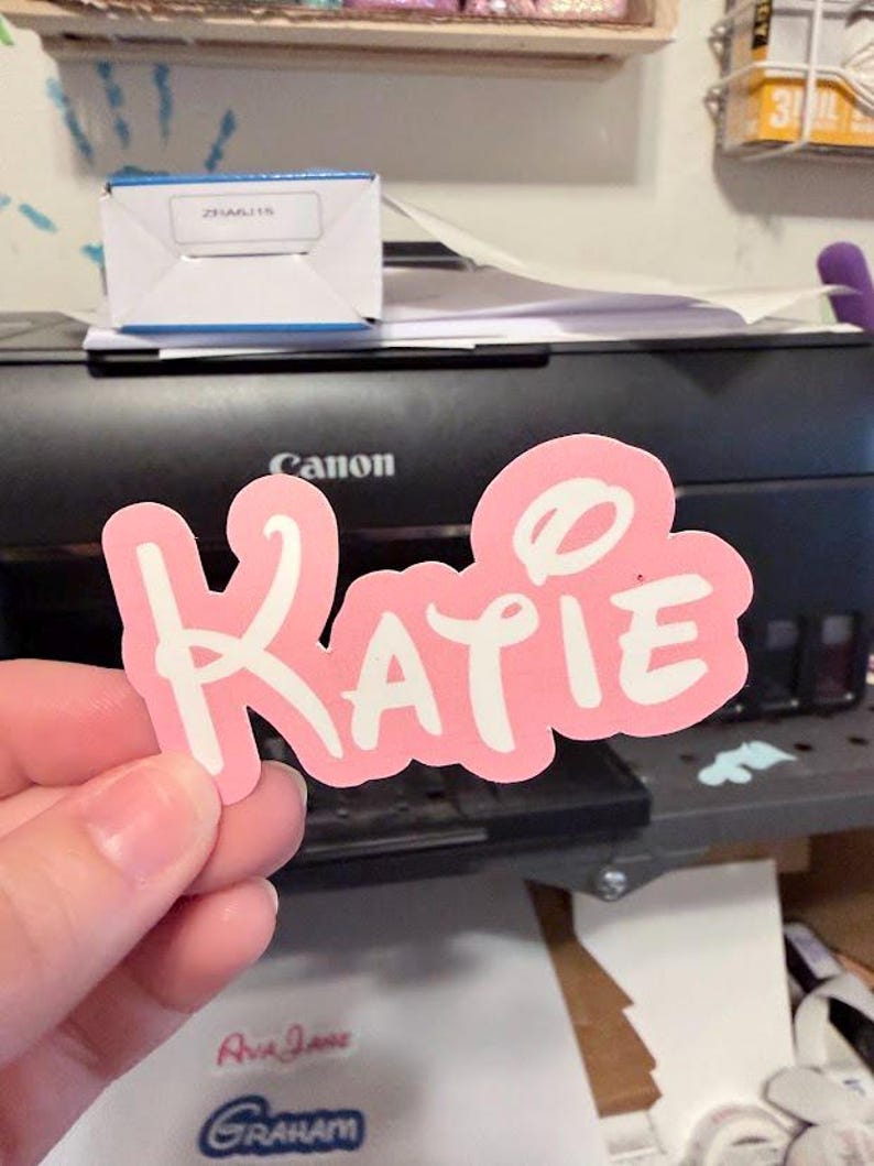 Peut inclure: Un autocollant rose avec le nom "Katie" en lettres blanches. L'autocollant a une bordure blanche et est tenu devant une imprimante noire. D'autres autocollants avec des noms sont visibles.