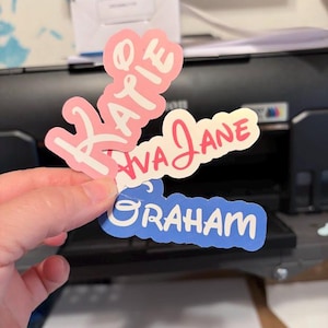 Peut inclure: Trois autocollants d&eacute;coup&eacute;s avec les noms "Katie" en rose, "Ava Jane" en rouge et "Graham" en bleu. Les autocollants sont tenus devant une imprimante.