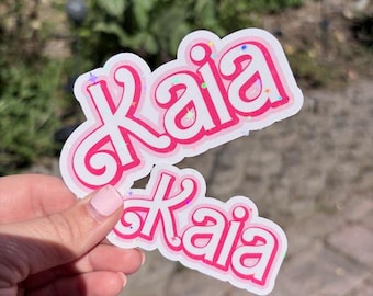 Könnte beinhalten: Zwei gestanzte Aufkleber mit dem Namen Kaia in einer fetten, rosa Schrift im Bubble-Stil mit weißem Rand. Die Schrift hat ein holografisches Finish, das kleine, bunte Stern- und Glitzerformen auf der Oberfläche des rosa Textes zeigt.