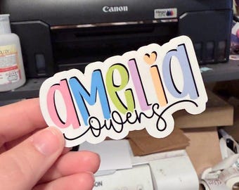 Könnte beinhalten: Ein gestanzter Aufkleber mit dem Namen Amelia in bunten, abgerundeten Blockbuchstaben mit einem kleinen Herz über dem Buchstaben i, und dem Namen Owens in schwarzer Schreibschrift darunter, alles auf weißem Hintergrund.