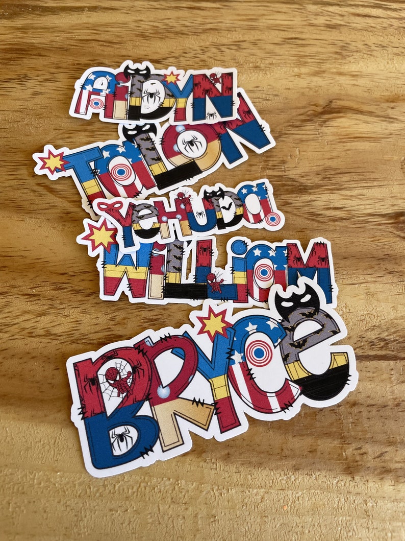 K&ouml;nnte beinhalten: F&uuml;nf personalisierte Namensaufkleber mit Superhelden-Motiven. Die Namen Aidyn, Talon, Yehuda, William und Bryce sind in fetten, bunten Buchstaben geschrieben, die mit Spinnennetzen, Fledermaussilhouetten und kreisf&ouml;rmigen Schildmotiven verziert sind.