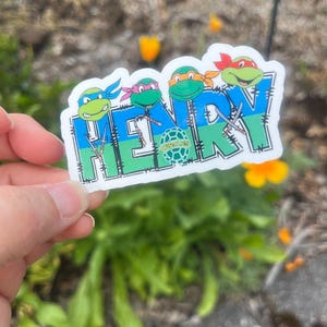 K&ouml;nnte beinhalten: Ein gestanzter Aufkleber mit dem Namen HENRY in gro&szlig;en blauen und gr&uuml;nen Blockbuchstaben. &Uuml;ber den Buchstaben befinden sich vier Zeichentrick-Schildkr&ouml;ten mit farbigen Masken, und kleine Ninja-Waffen sind in das Design integriert.