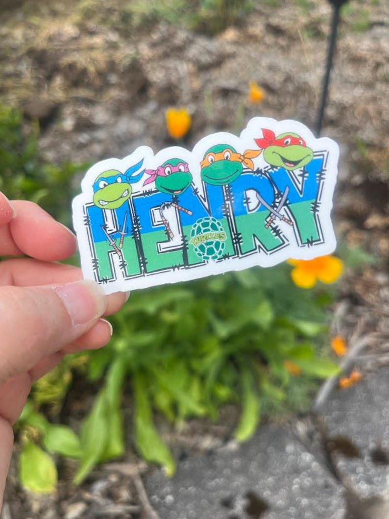 K&ouml;nnte beinhalten: Ein gestanzter Aufkleber mit dem Namen HENRY in gro&szlig;en blauen und gr&uuml;nen Blockbuchstaben. &Uuml;ber den Buchstaben befinden sich vier Zeichentrick-Schildkr&ouml;ten mit farbigen Masken, und kleine Ninja-Waffen sind in das Design integriert.